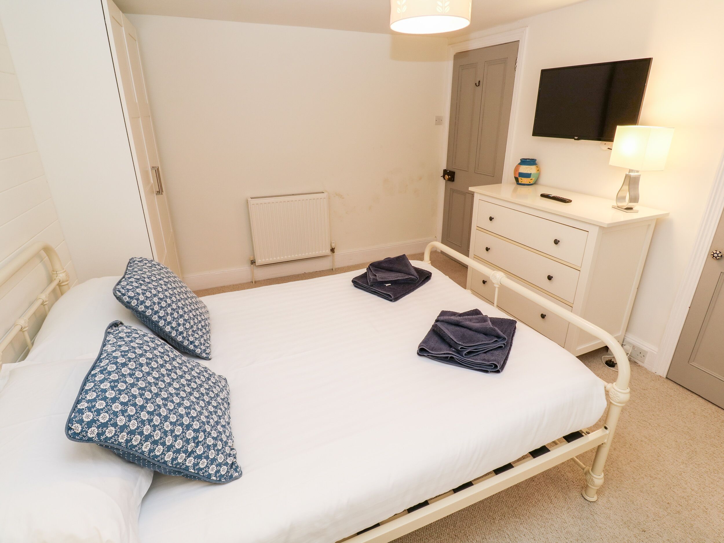 Un lit bien fait, avec des oreillers et des serviettes pliées, une table de chevet avec une lampe, une commode, un radiateur et un téléviseur fixé au mur.