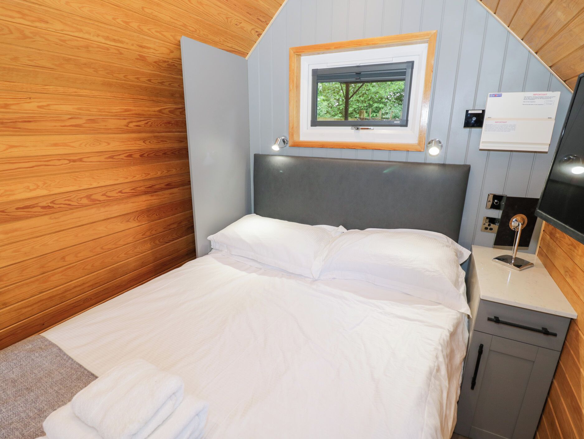 A neatly made bed with white linens, a wooden headboard, a small window with a view of trees, and a bedside table with a lamp and a note.