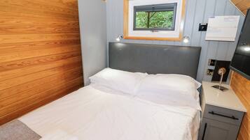 A neatly made bed with white linens, a wooden headboard, a small window with a view of trees, and a bedside table with a lamp and a note.