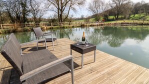Een houten terras met een lounge stoel en een tafel, een champagnefles en uitzicht op een rustig meer met bomen op de achtergrond.