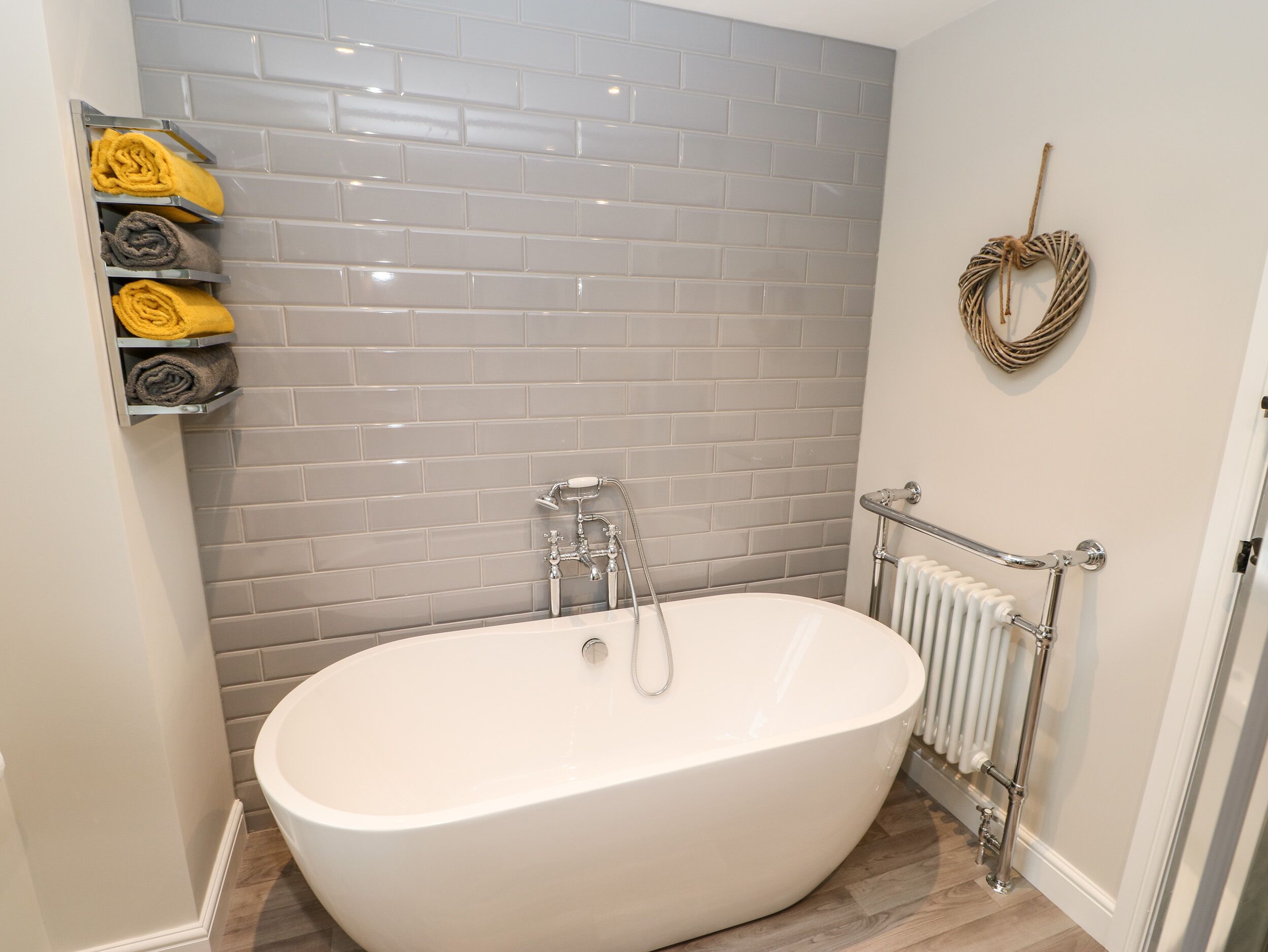 A modern bathroom with a freestanding bathtub, grey tiled walls, a towel rack with folded towels, a radiator, and a decorative wreath.