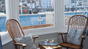 Two wooden chairs with cushions, a small table with glasses and a tray, and a view of a blue pool and buildings through a window. - Seasides (Weymouth)