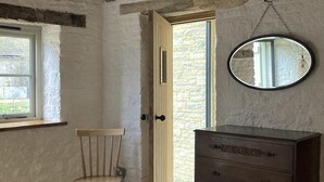 Una stanza con una sedia di legno, un comò, uno specchio e una finestra vista su un muro in pietra.
