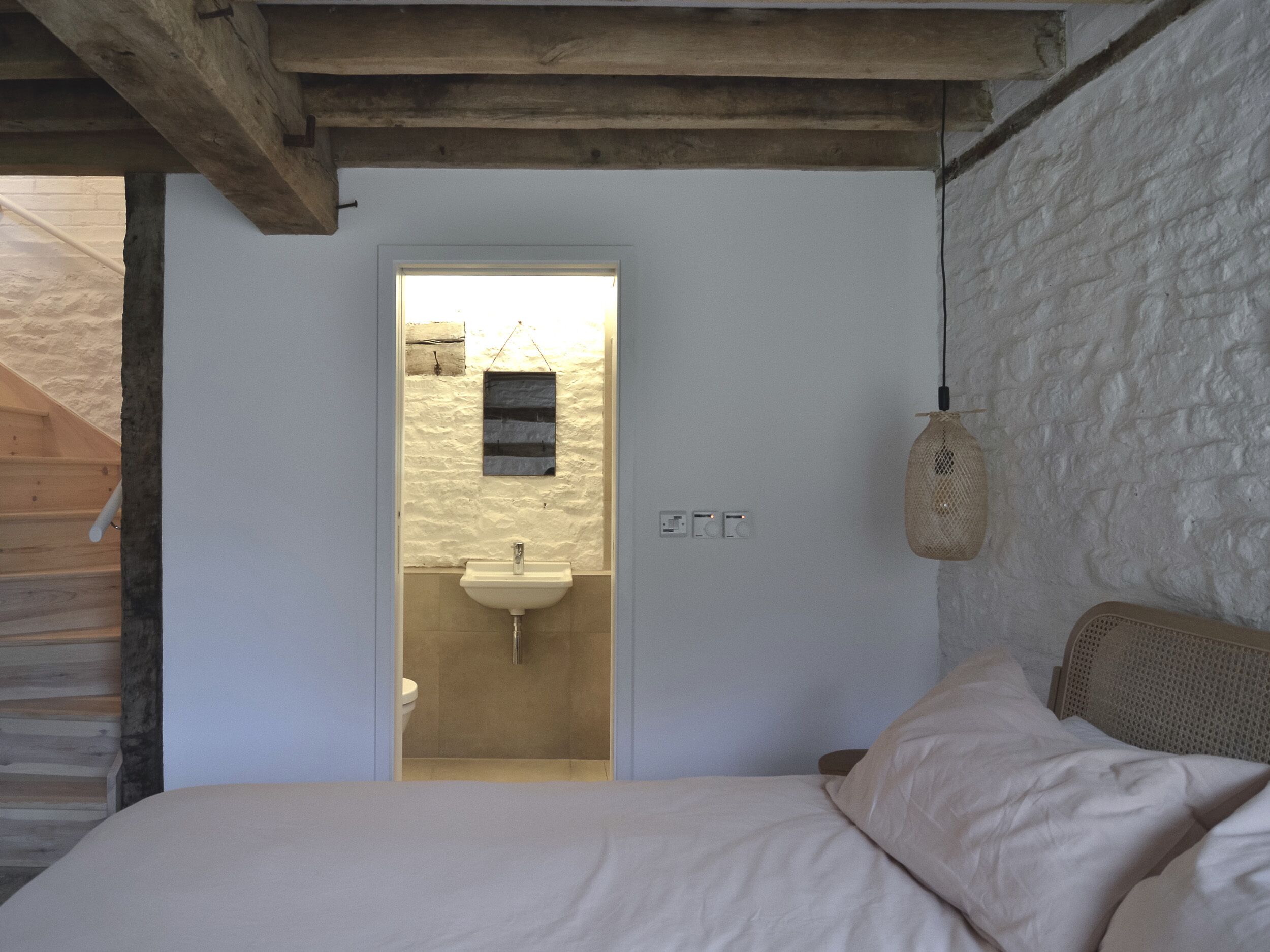 A bedroom with a wooden ceiling, a bed with white bedding, a hanging light fixture, and a small window with a view of a bathroom.