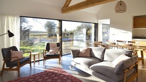 A spacious living room with a large window, a sofa, a chair, and a dining area with wooden furniture. - High Cogges Farm Holiday Cottages The Cart Shed (Witney)