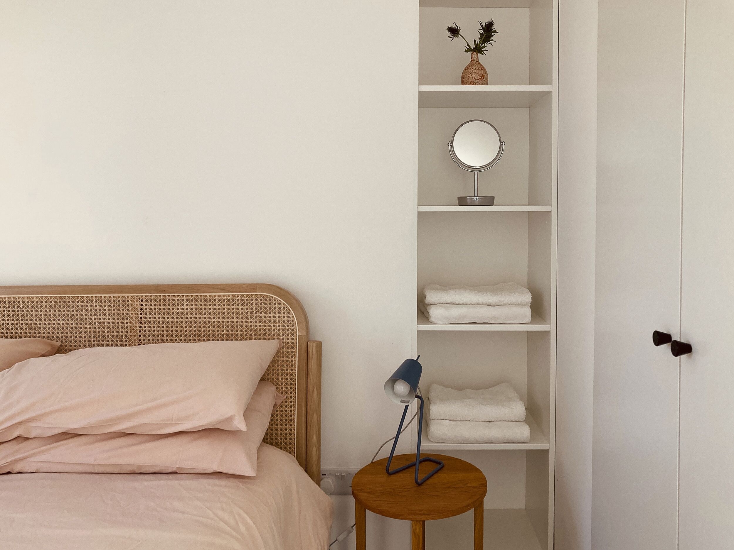 A bedroom with a bed, a bedside table with a lamp, a shelf with towels, a mirror, and a vase with flowers.