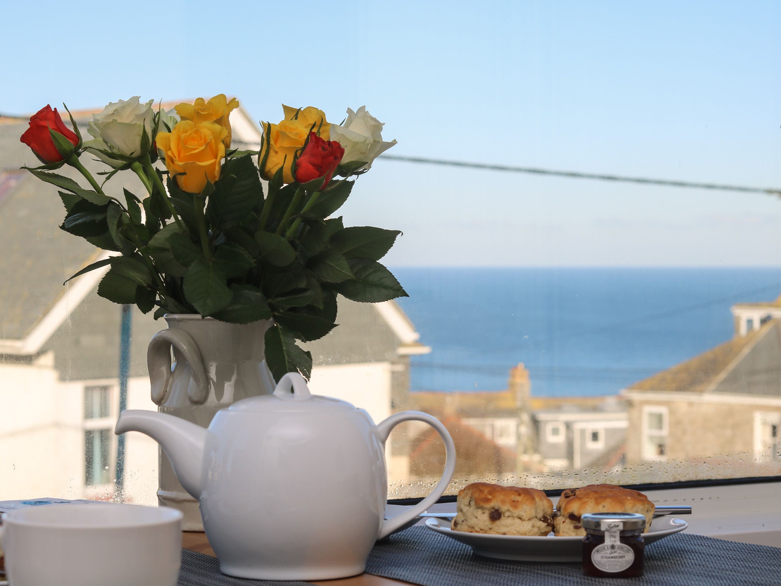 A table with a white teapot, a cup, a plate with biscuits, and a vase with roses.