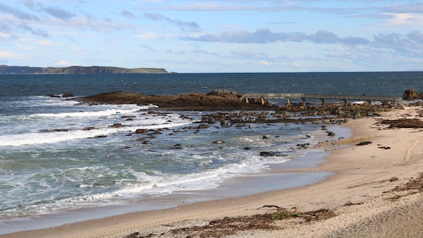 Una playa rocosa con olas rompiendo en la orilla, un muelle de madera y acantilados distantes bajo un cielo parcialmente nublado.