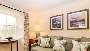 A living room with a green sofa, floral cushions, a table with a bowl, a lamp, and two framed pictures on the wall. - South Mains Cottage - Craigievar Castle (Alford)