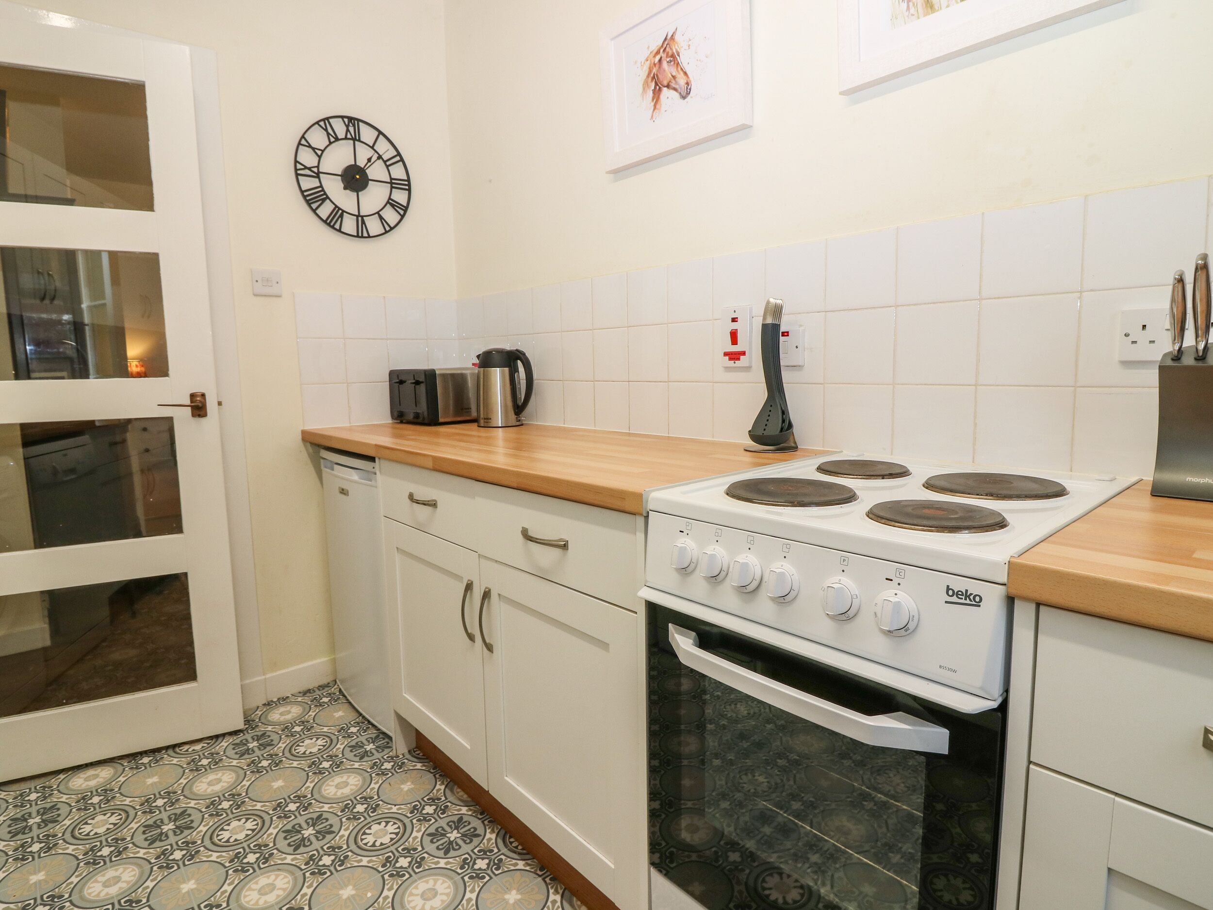 Une cuisine avec des meubles blancs, une cuisinière Beko, un grille-pain et une horloge au mur.