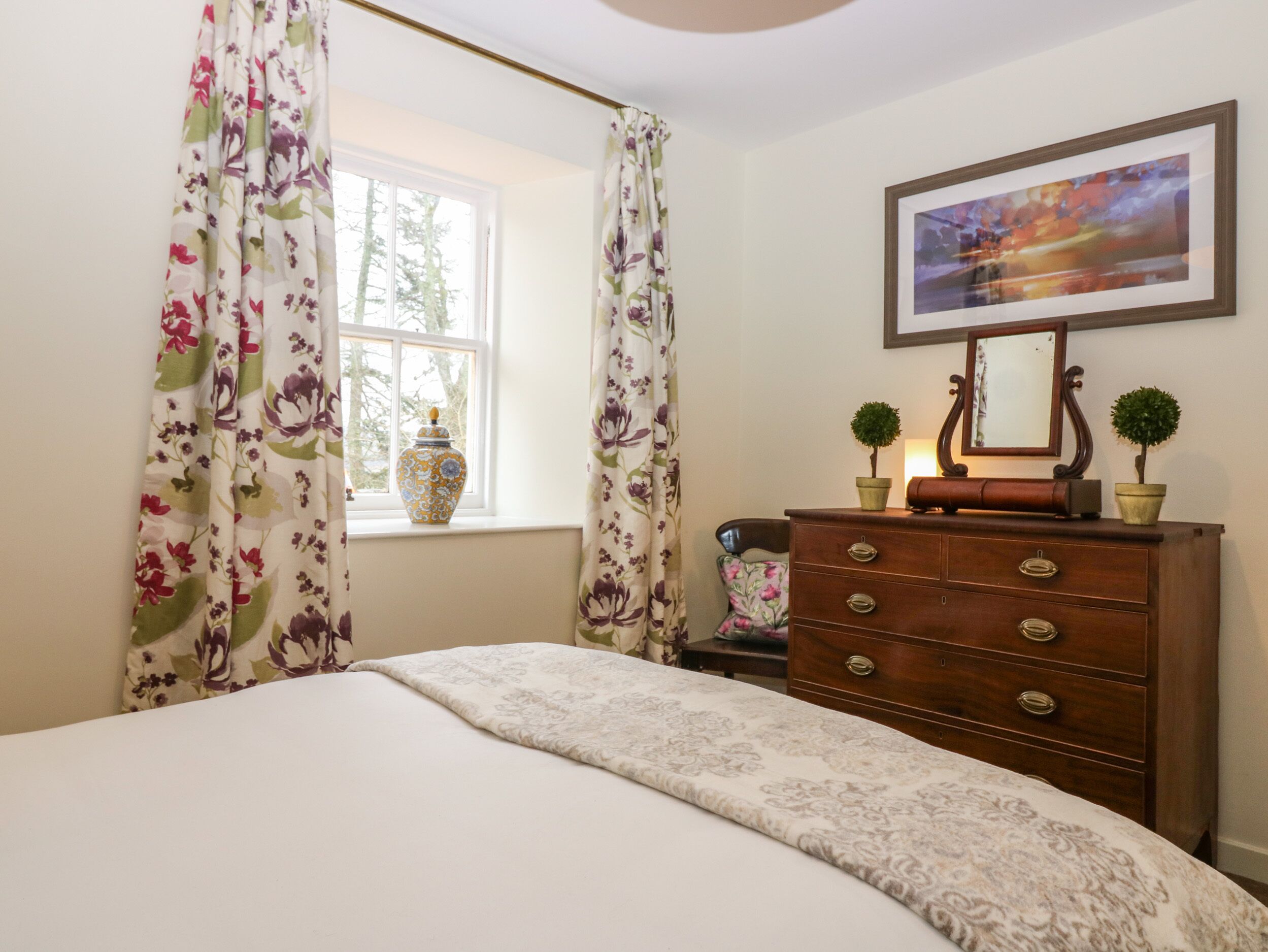 A bedroom with a bed, a dresser, a window with curtains, a vase, and a framed picture on the wall.