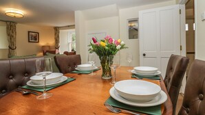 A dining room with a wooden table set for four, surrounded by leather chairs. A vase with flowers is placed in the center.