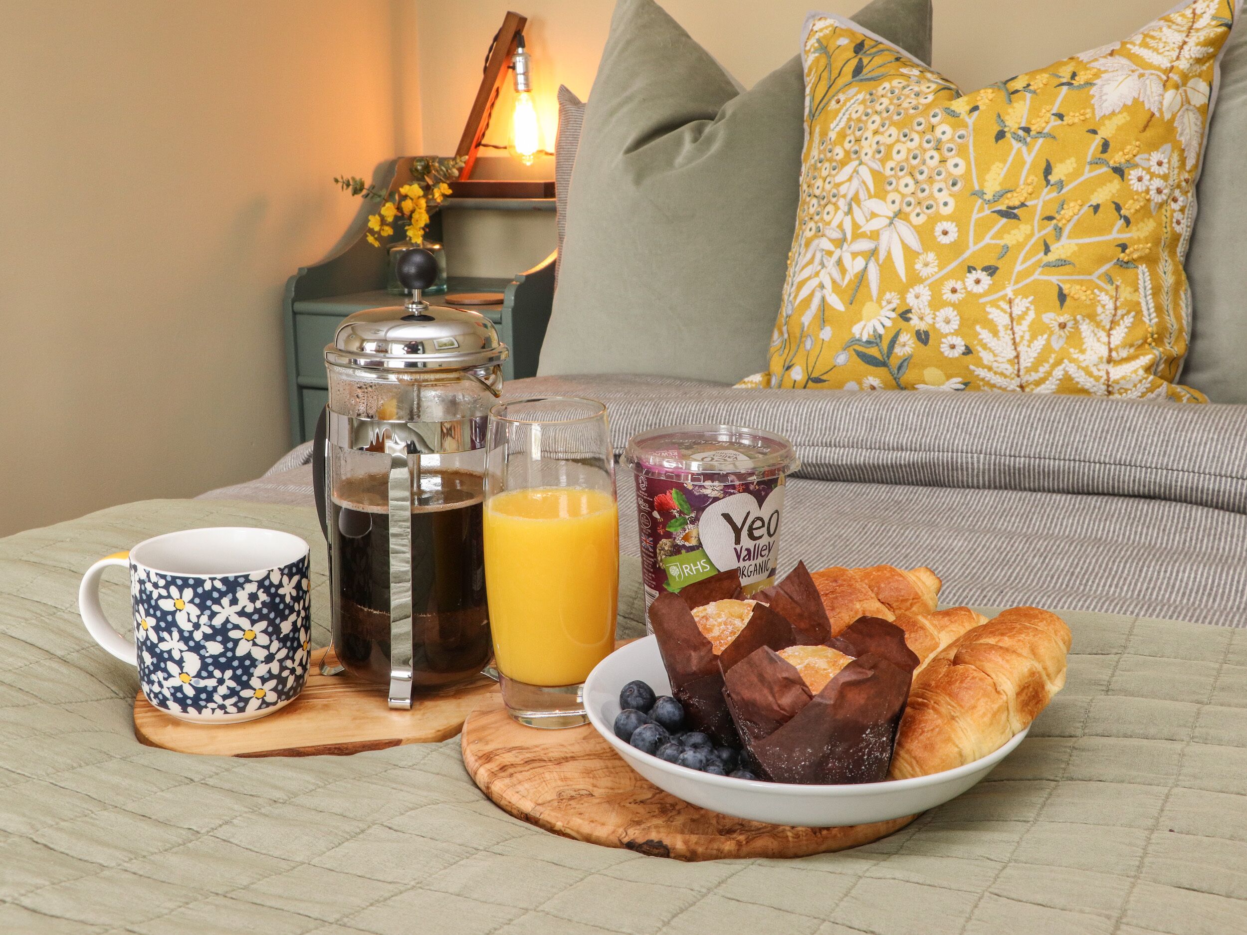 A breakfast setup with a coffee pot, mug, glass of orange juice, and a bowl of croissants and blueberries on a wooden board.