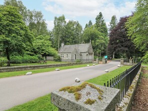 Un bâtiment en pierre à l’architecture traditionnelle, entouré d’une végétation luxuriante et bordé d’une route pavée.