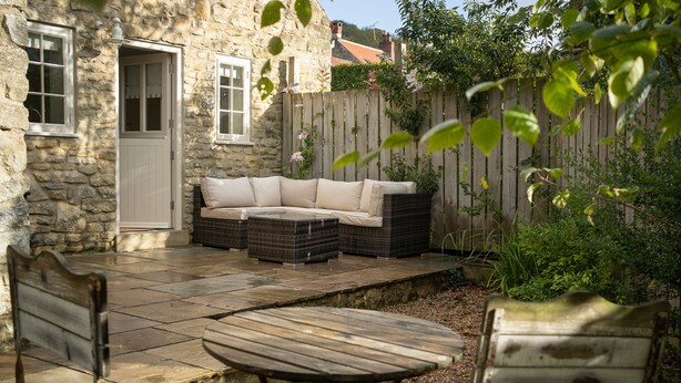 Une maison en pierre avec une terrasse en bois, une porte blanche, un canapé beige et une table en bois.