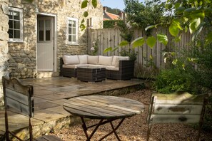 Une maison en pierre avec une terrasse en bois, une porte blanche, un canapé beige et une table en bois.