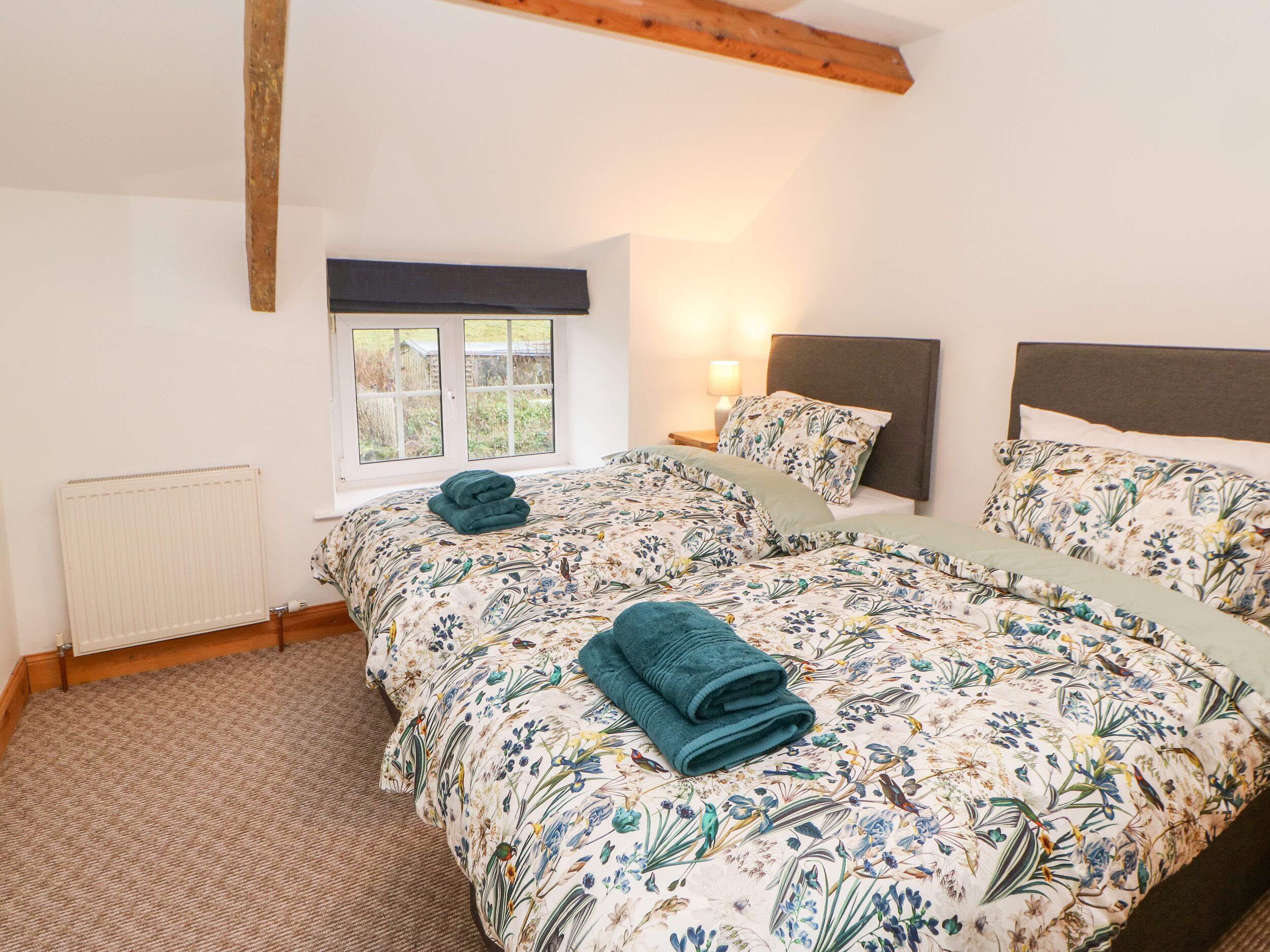 Un dormitorio con dos camas, ropa de cama floral, una ventana con vistas, un radiador y una viga de techo de madera.