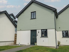 Et moderne hus i to etager med grøn facade og hvide vægge, med en hoveddør og en lille veranda samt en potteplante ved siden af.