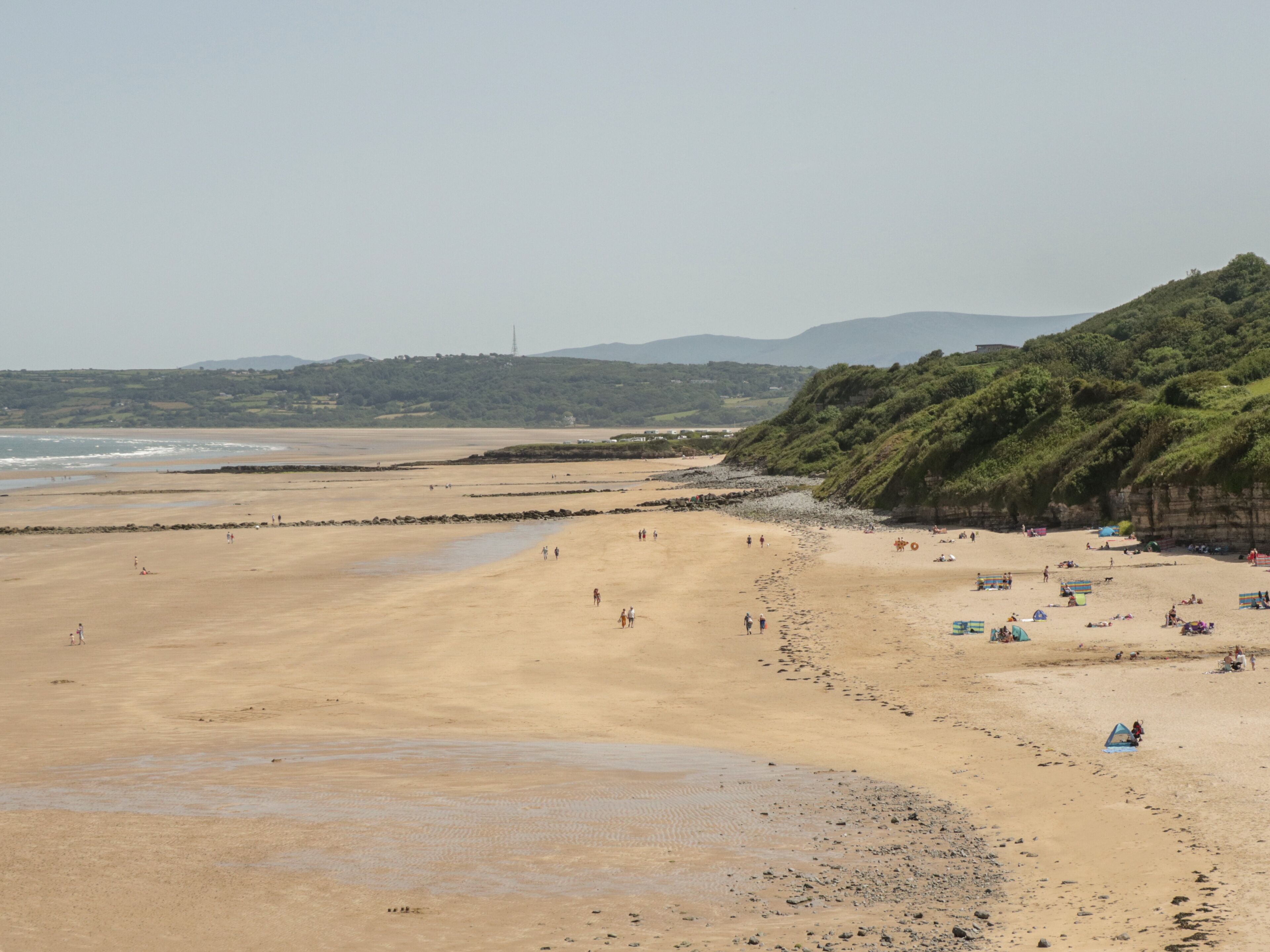 A wide sandy beach with people and tents, a rocky shoreline, and hills in the background.