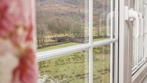 Vista a través de una ventana que muestra un muro de piedra y árboles, con montañas al fondo.