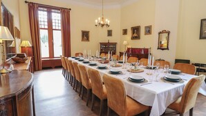 Een elegante eetzaal met een lange tafel gedekt voor een maaltijd, houten stoelen, een kroonluchter en kunstwerken in lijsten aan de muren.