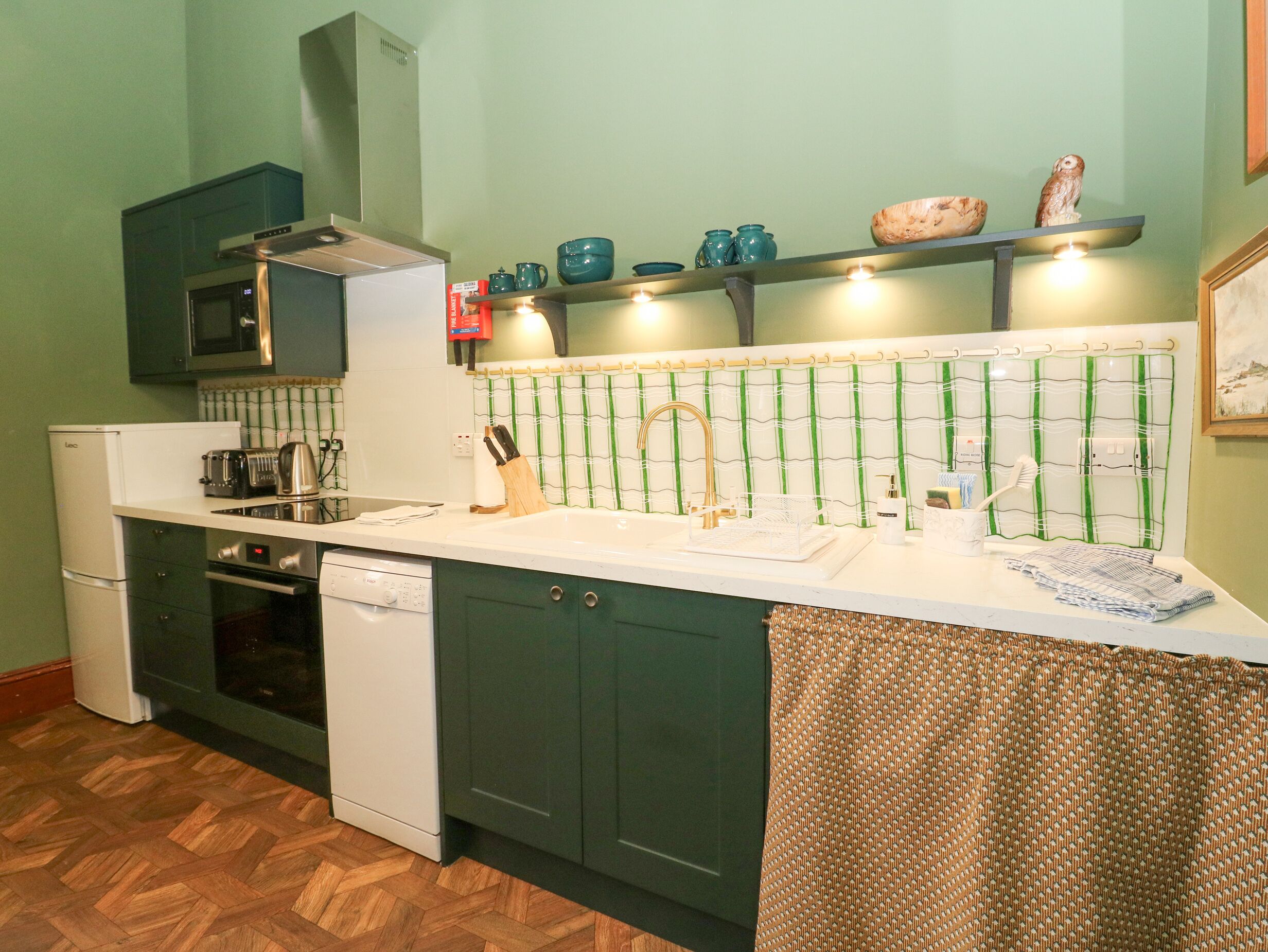 A compact kitchen with a microwave, oven, and dishwasher. There is a sink with a faucet, and a shelf above it holding various kitchen items.