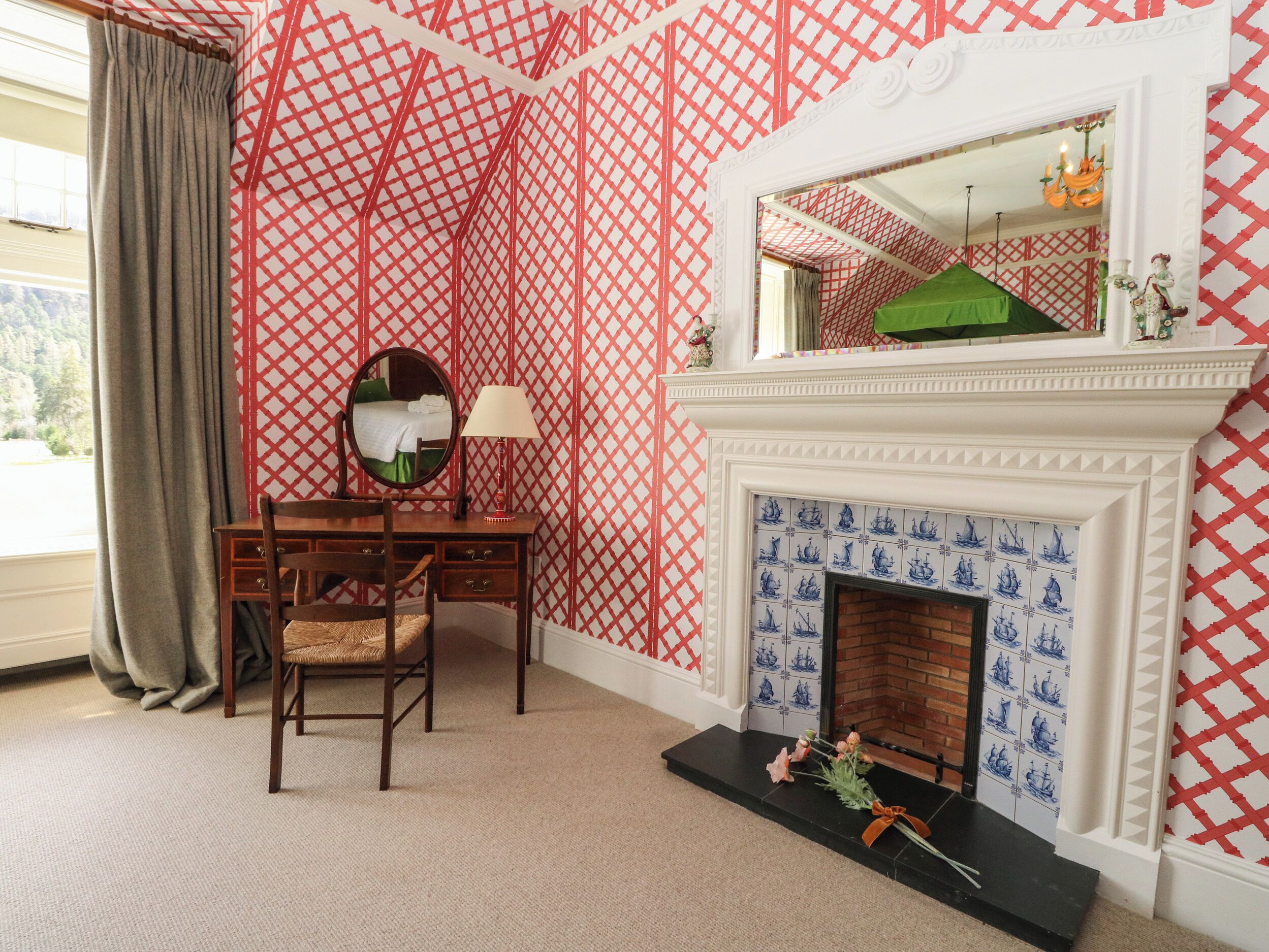 A room with a patterned red and white wallpaper, a wooden desk with a mirror, a lamp, and a fireplace with blue tiles.