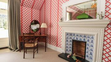 A room with a patterned red and white wallpaper, a wooden desk with a mirror, a lamp, and a fireplace with blue tiles.