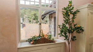 Una habitación con ventanal panorámico que da a un muro de ladrillo y árboles, un banco blanco con una cesta de flores y una planta en maceta con hojas amarillas.