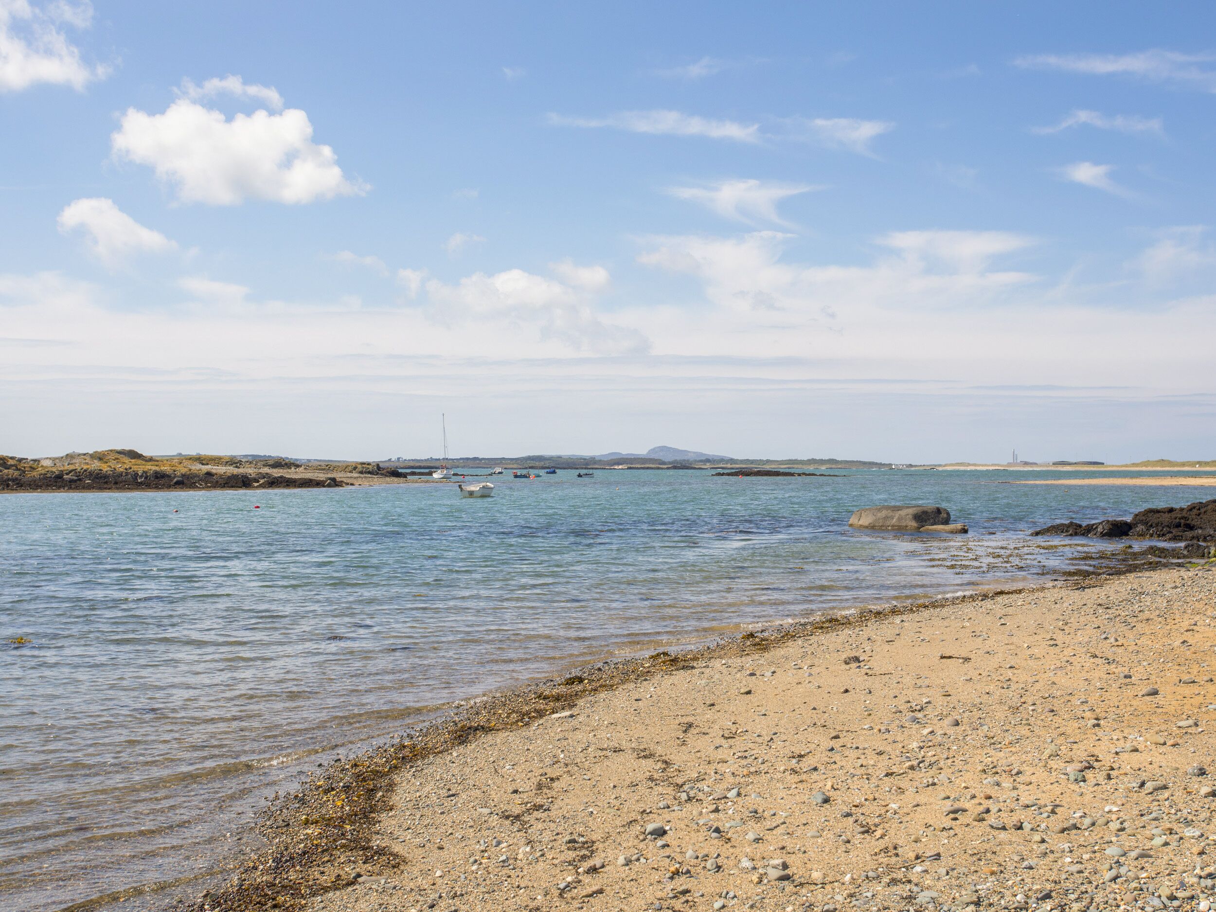 A coastal beach with calm waters, pebbly shore, and distant hills.