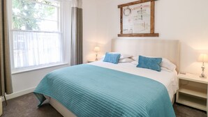A neatly arranged bedroom with a large bed, two bedside tables, a window with curtains, and a wall-mounted artwork. - Babbacombe Hall (Torquay)