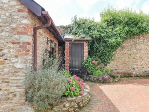 A stone building with a small wooden door, a brick pathway, and a garden with pink flowers. - Coach Cottage (Colyton)