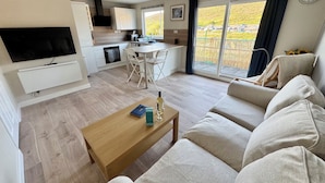 A modern living room with a sofa, a wooden coffee table, a TV, and a kitchen area with a dining table. - Gecko Beach (Truro)