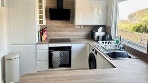 A compact kitchen with a built-in oven, microwave, and sink, a window with a view of greenery, and a black range hood. - Gecko Beach (Truro)