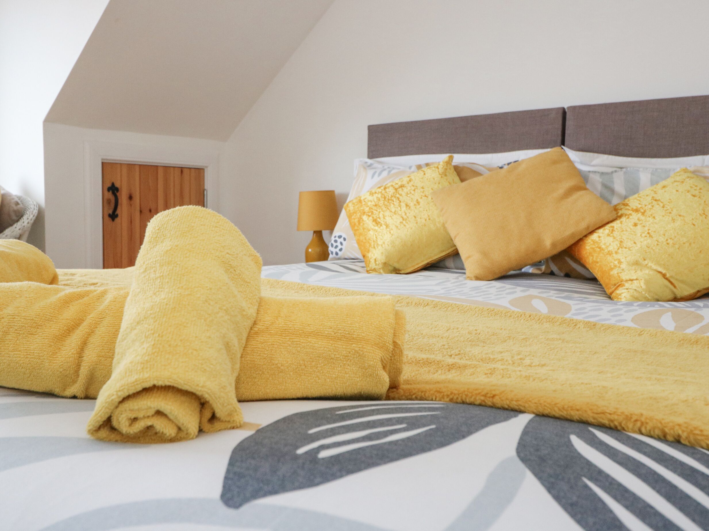 A neatly made bed with yellow linens and pillows, a wooden door, and a lamp on a bedside table.