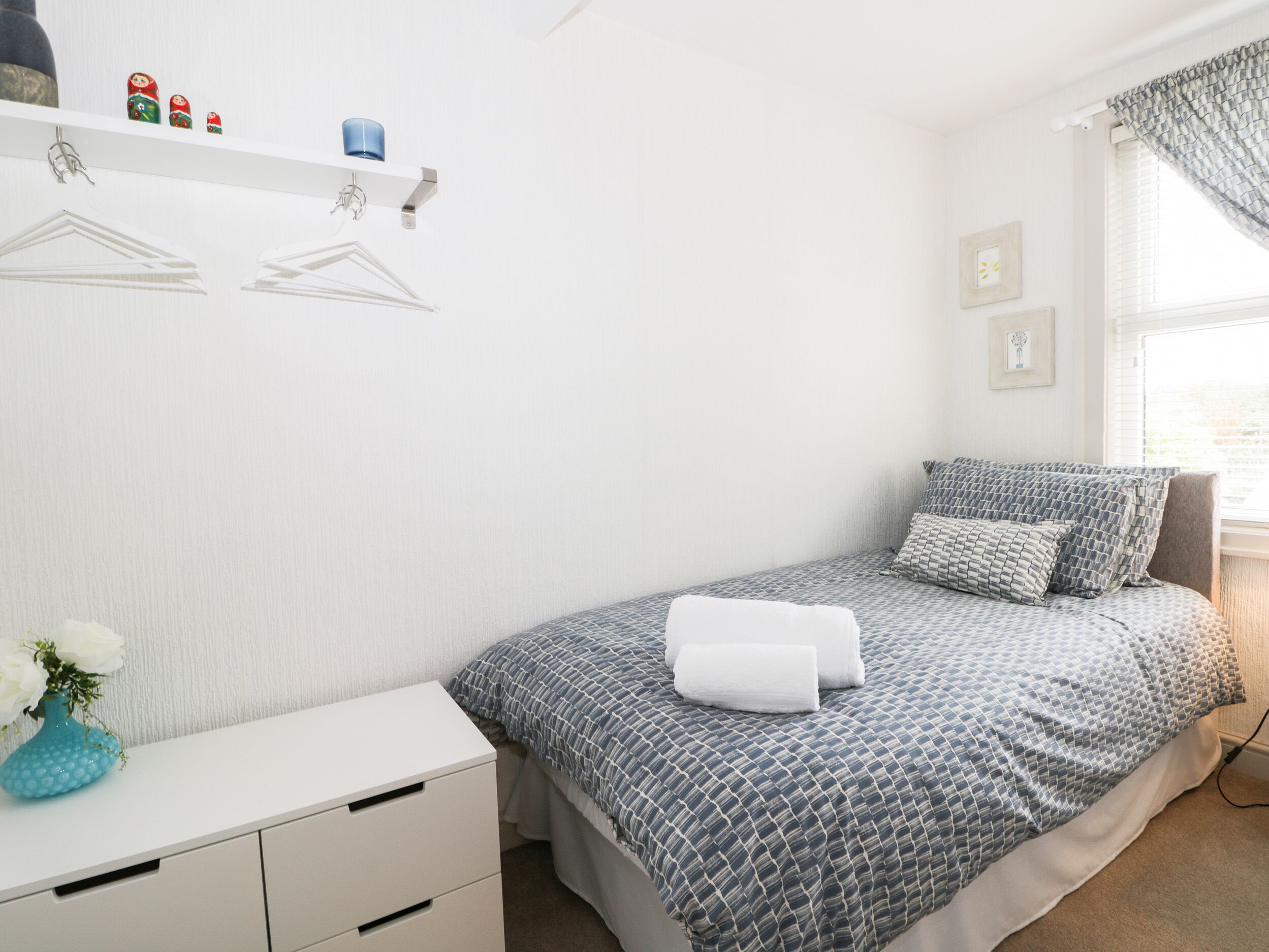 A bedroom with a bed, a dresser, a shelf with decorative items, and a window with curtains.