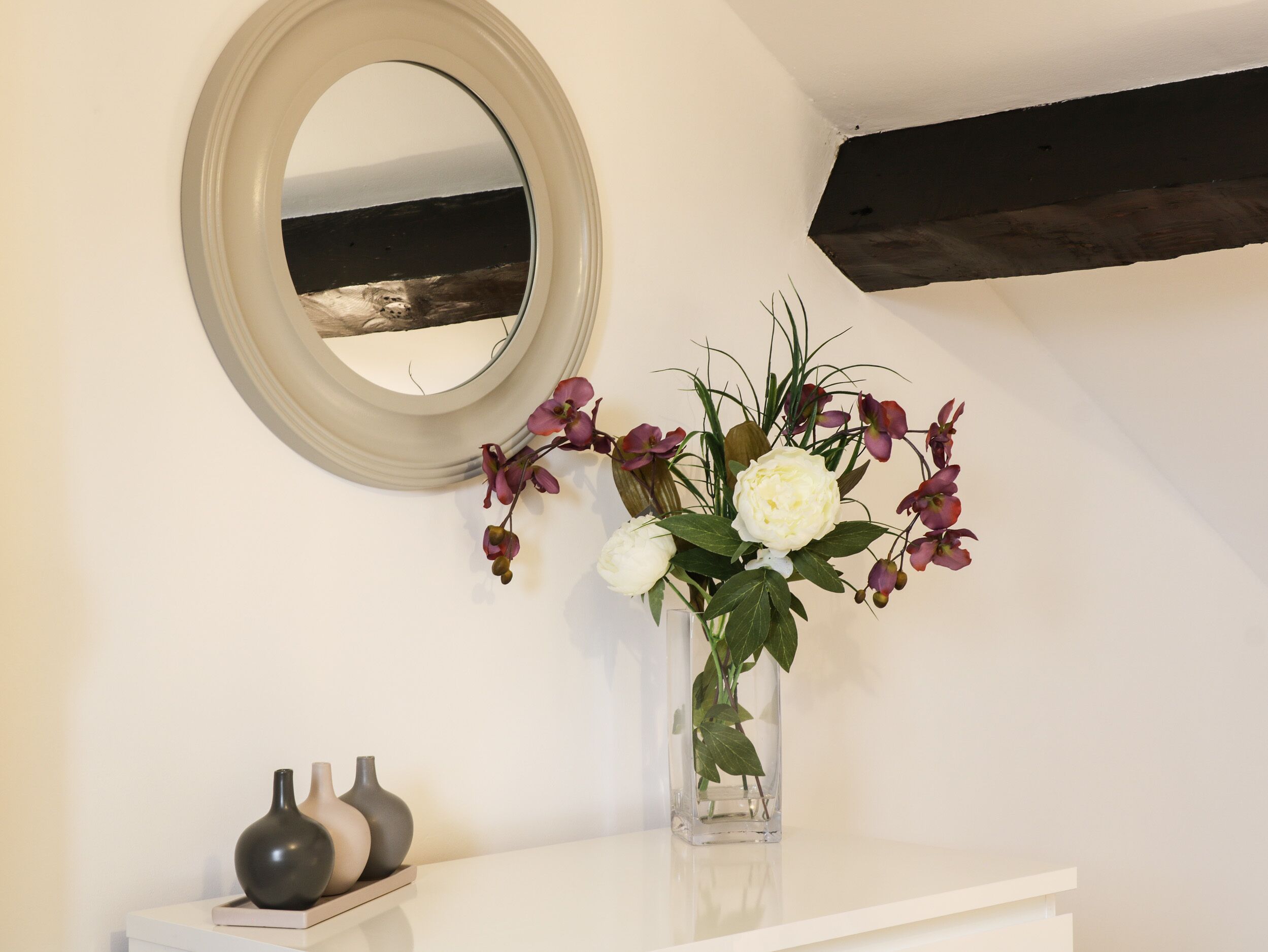 Une salle de bain avec un meuble-lavabo blanc, un miroir rond et un vase rempli de fleurs.