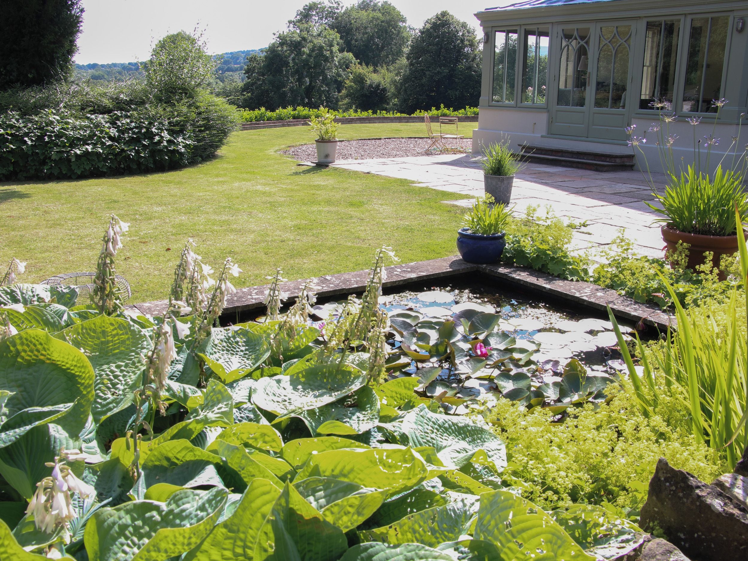 A garden with a pond, potted plants, and a conservatory.