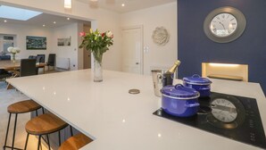 A modern kitchen with a central island, pendant lighting, a clock, and a vase with flowers. - Greenlawns (Burnham-on-Sea)