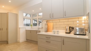A modern kitchen with white cabinets, a countertop with a kettle, toaster, and cups, and a window with a view of the outside. - Greenlawns (Burnham-on-Sea)
