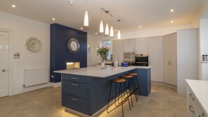 A modern kitchen with a central island, pendant lights, a clock, and a radiator. - Greenlawns (Burnham-on-Sea)