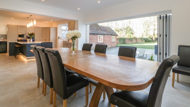 A modern kitchen with a central island, a dining area with a large wooden table and black chairs, and a view of a brick house through a sliding glass door. - Greenlawns (Burnham-on-Sea)