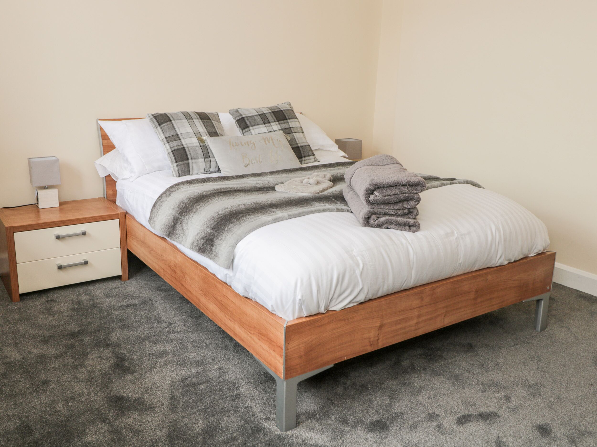 Een net opgemaakt bed met een grijs-wit gestreept dekbed, een opgevouwen grijze handdoek en ruitkussens. Er staat een nachtkastje met een lamp en een klein doosje. De kamer heeft een vloerbedekking en een effen muur.