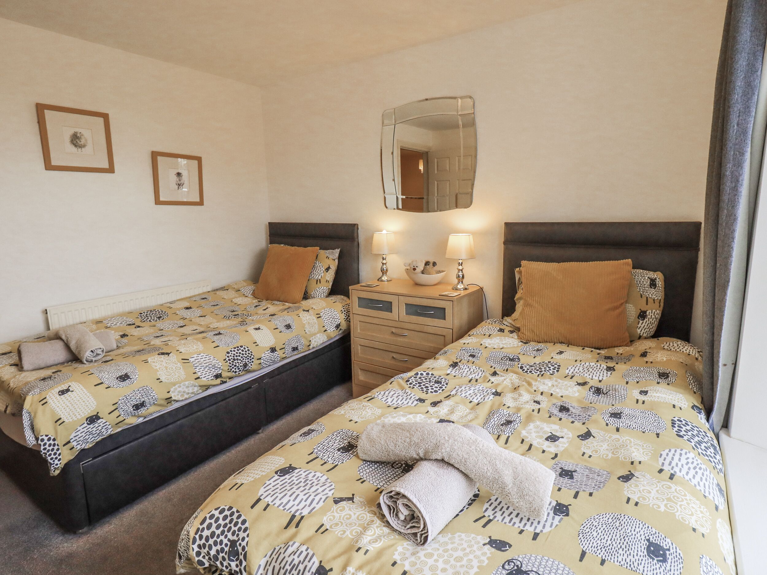 A double room with two beds, each with a patterned bedspread, a bedside table with a mirror, and two wall-mounted pictures.