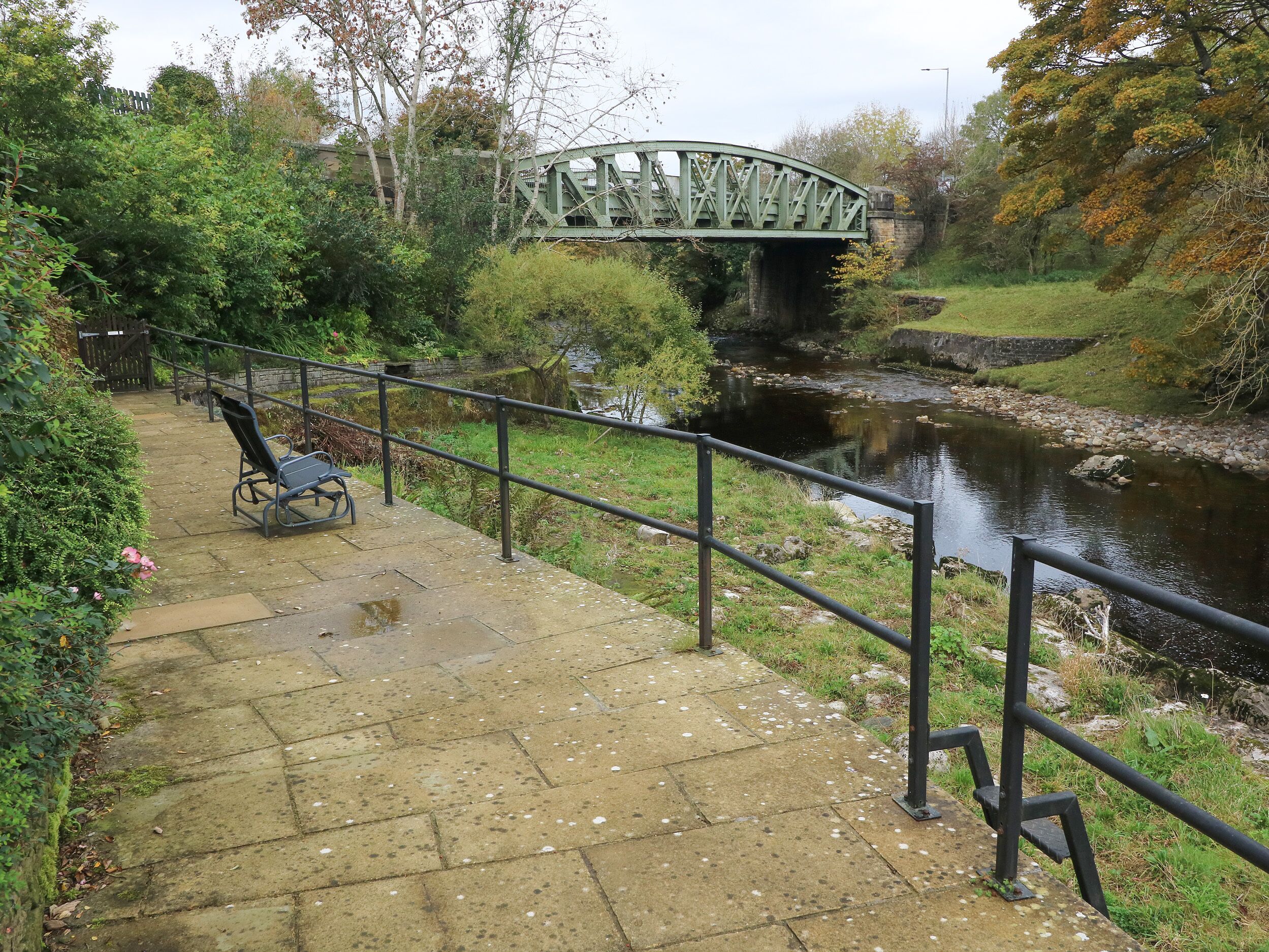 A stone pathway leading to a bench overlooking a river, with a green bridge in the background.