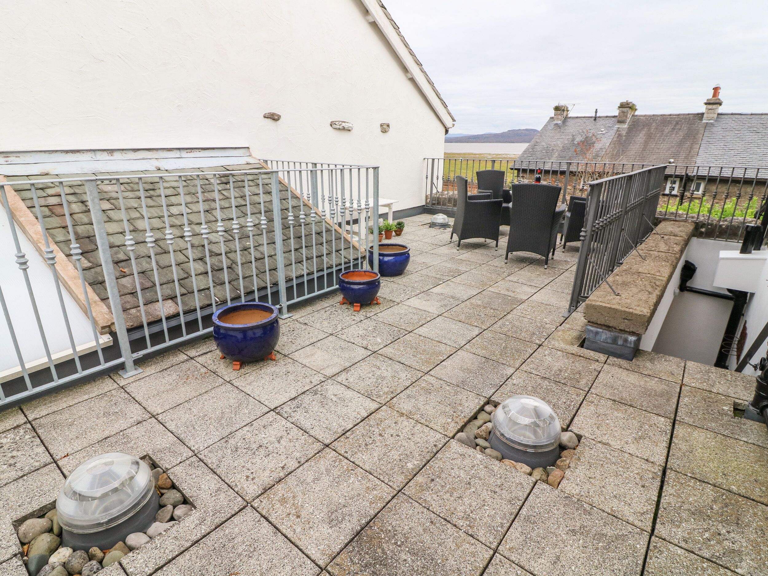 Um terraço no telhado com azulejos de pedra, dois vasos azuis, um lareira com cobertura de vidro e uma cerca de metal.