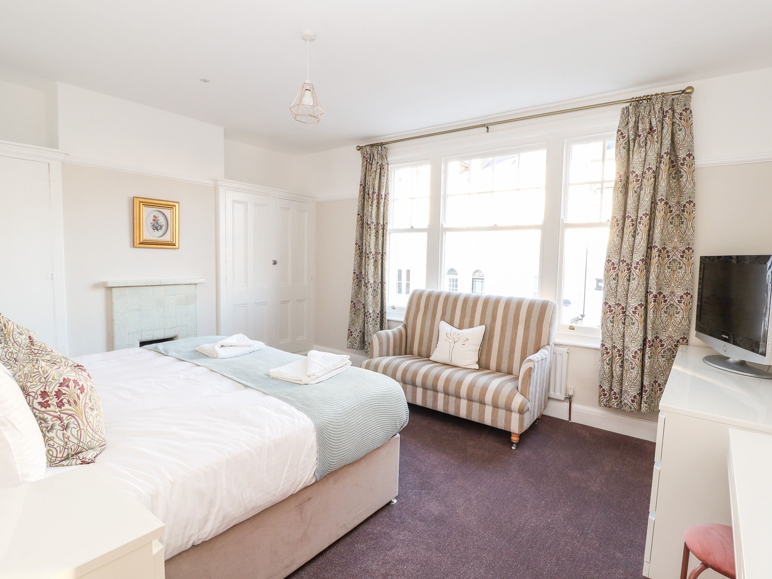 A hotel room with a large bed, a striped armchair, a desk with a television, and a window with curtains.