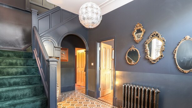 A staircase with green carpet leading to a room with patterned flooring and a radiator. - Livingstone House (Llandudno)
