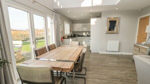 A spacious dining area with a wooden table and chairs, a kitchen in the background, and large windows offering a view of the outdoors. - The Deck House (Chathill)