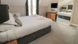 A modern bedroom with a large bed, a dresser, a TV, and a sliding glass door leading to a balcony. - The Deck House (Chathill)